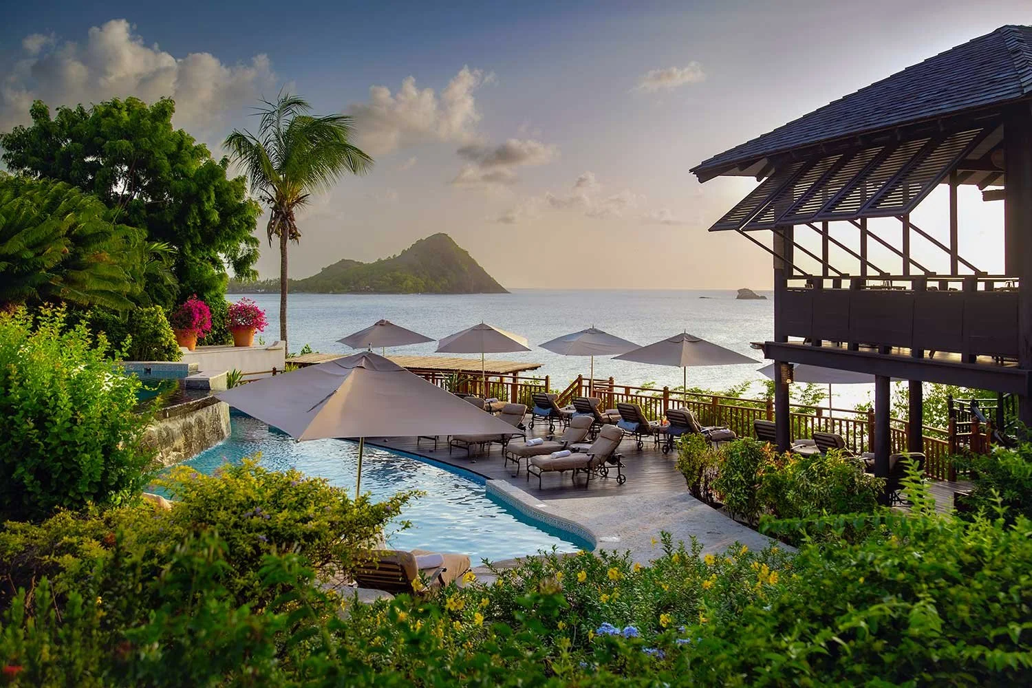 Luxury resort infinity pool in St. Lucia surrounded by lush tropical gardens, oceanfront lounge chairs, and stunning Caribbean Sea views at sunset.