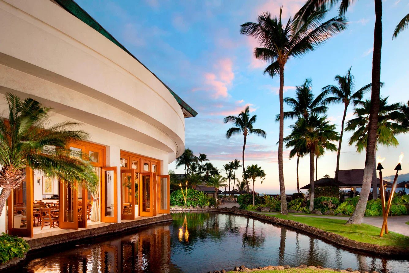 Beautiful tropical resort in Maui at sunset with palm trees, lagoon reflecting the sky, and open-air dining overlooking lush gardens.