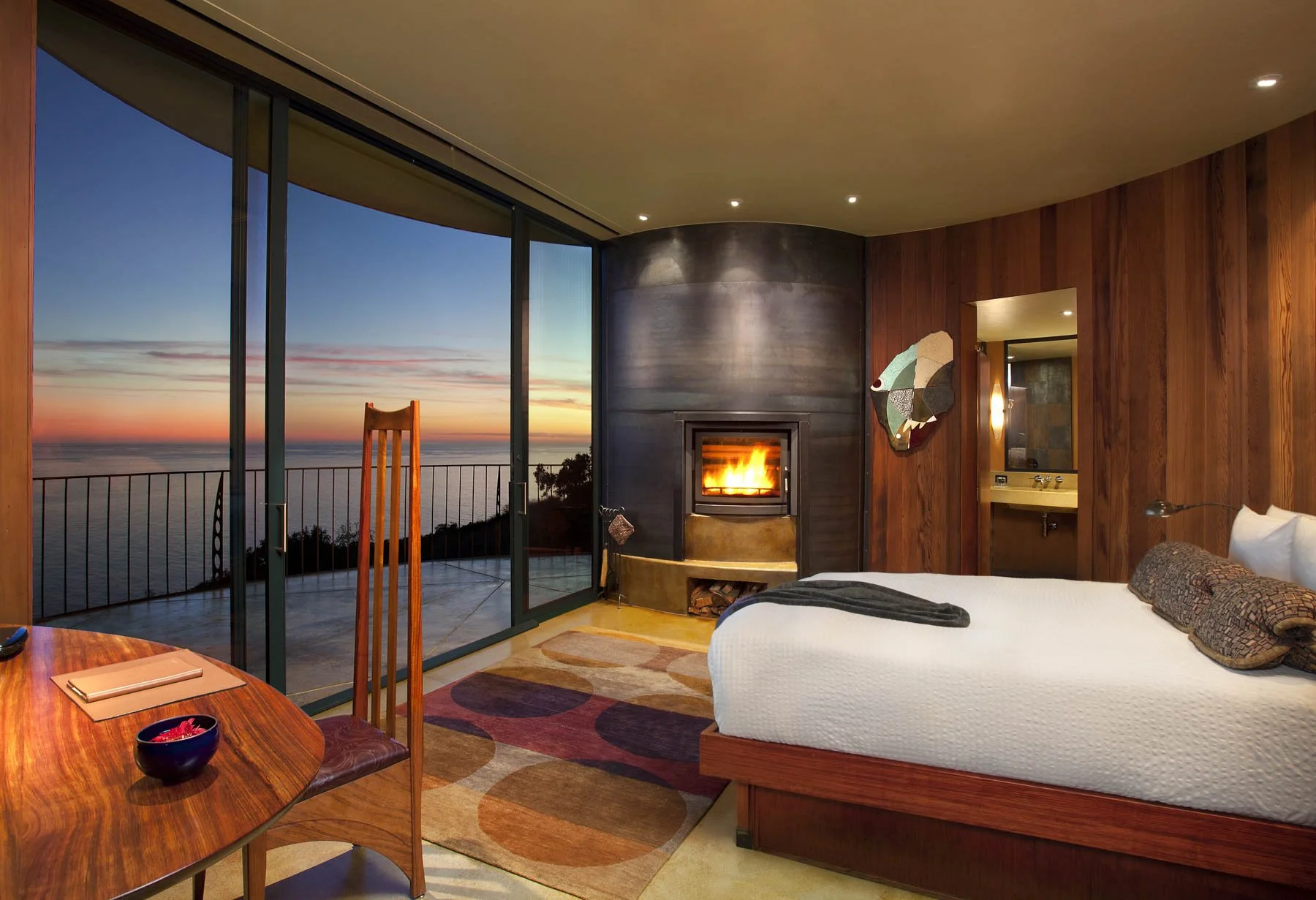 Luxury guest room at Post Ranch Inn in Big Sur, California, featuring warm wood interiors, a fireplace, and floor-to-ceiling windows overlooking the Pacific Ocean at sunset.