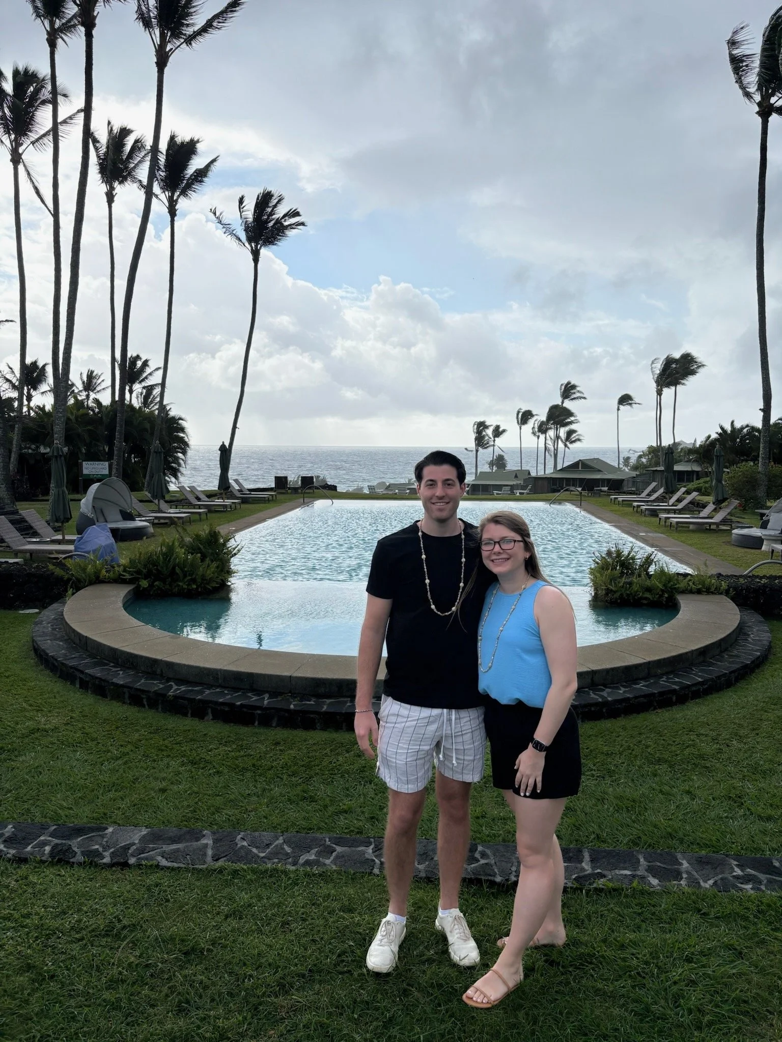 Couple standing in front of the oceanfront infinity pool at Hana-Maui Resort with palm trees and Pacific Ocean views on Maui’s east coast.