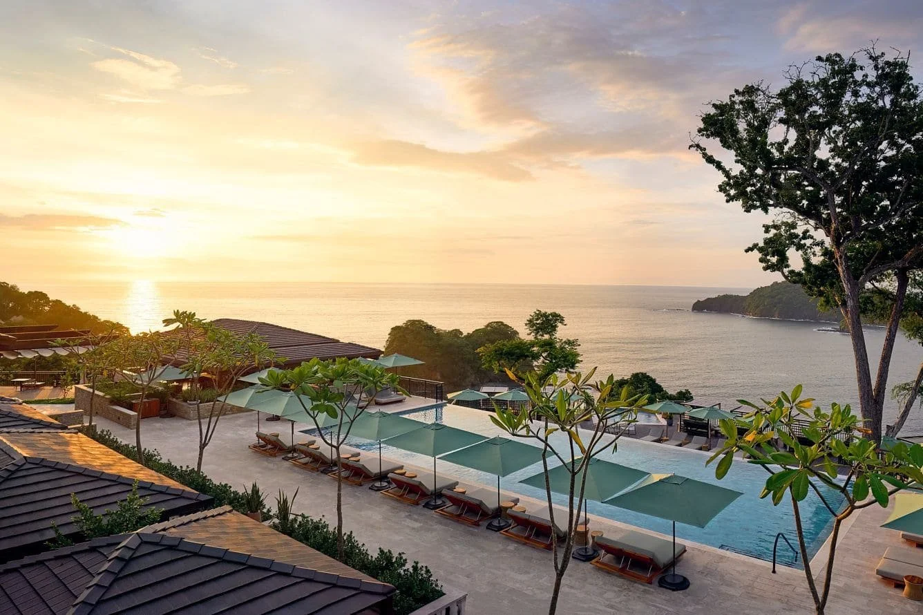 Infinity pool overlooking the Pacific Ocean at Nekajui, a Ritz-Carlton Reserve in Costa Rica, with lounge chairs, tropical landscaping, and a glowing sunset illuminating the coastline.