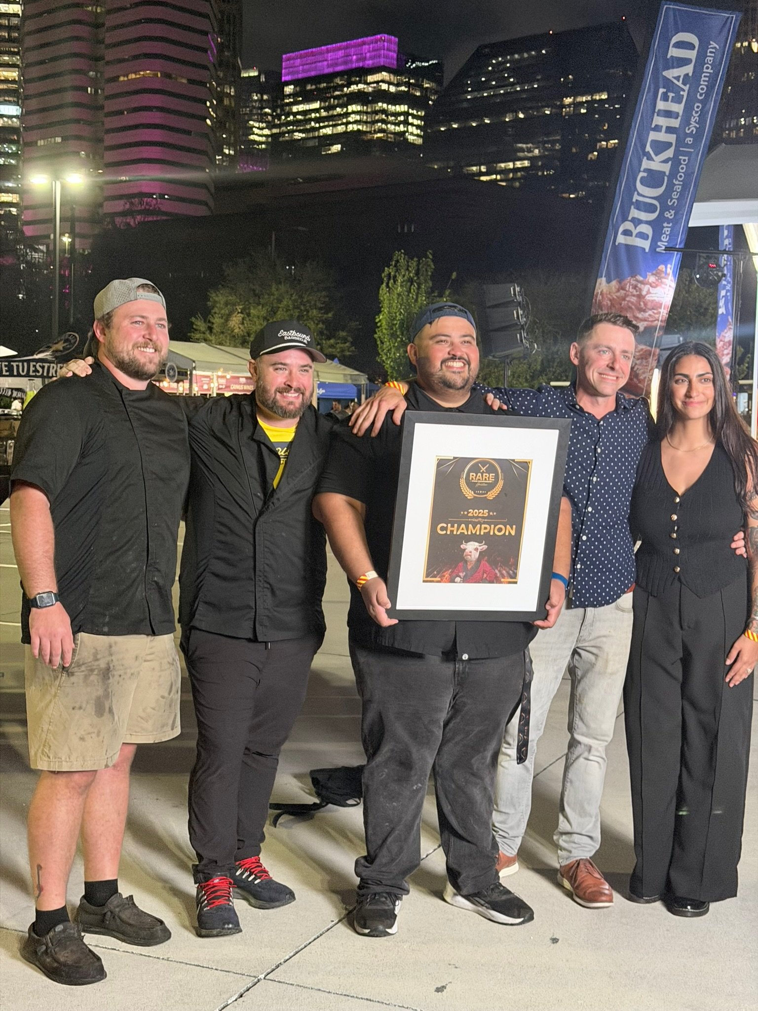 A group of five people standing outdoors at night, celebrating and smiling. One person in the center is holding a framed certificate that says '2025 CHAMPION' with a graphic of a bull and the word 'RARE'. The background features tall buildings with colorful lights and a large banner that reads 'BUCKHEAD'.