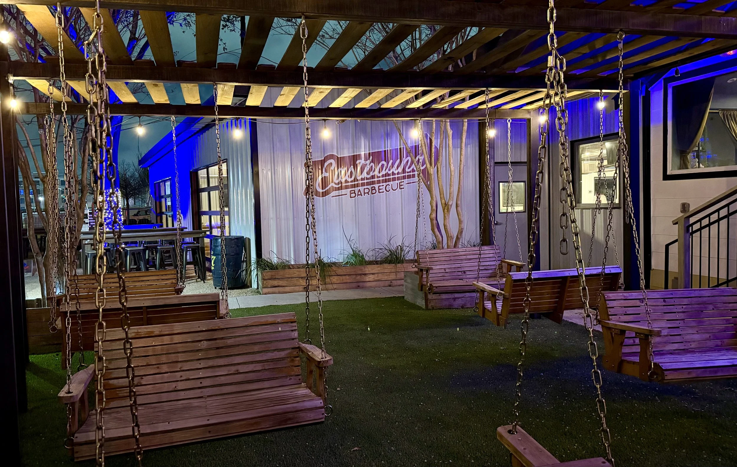 Outdoor seating area with wooden swings hanging from a pergola, string lights, and a sign for courtyard barbecue.