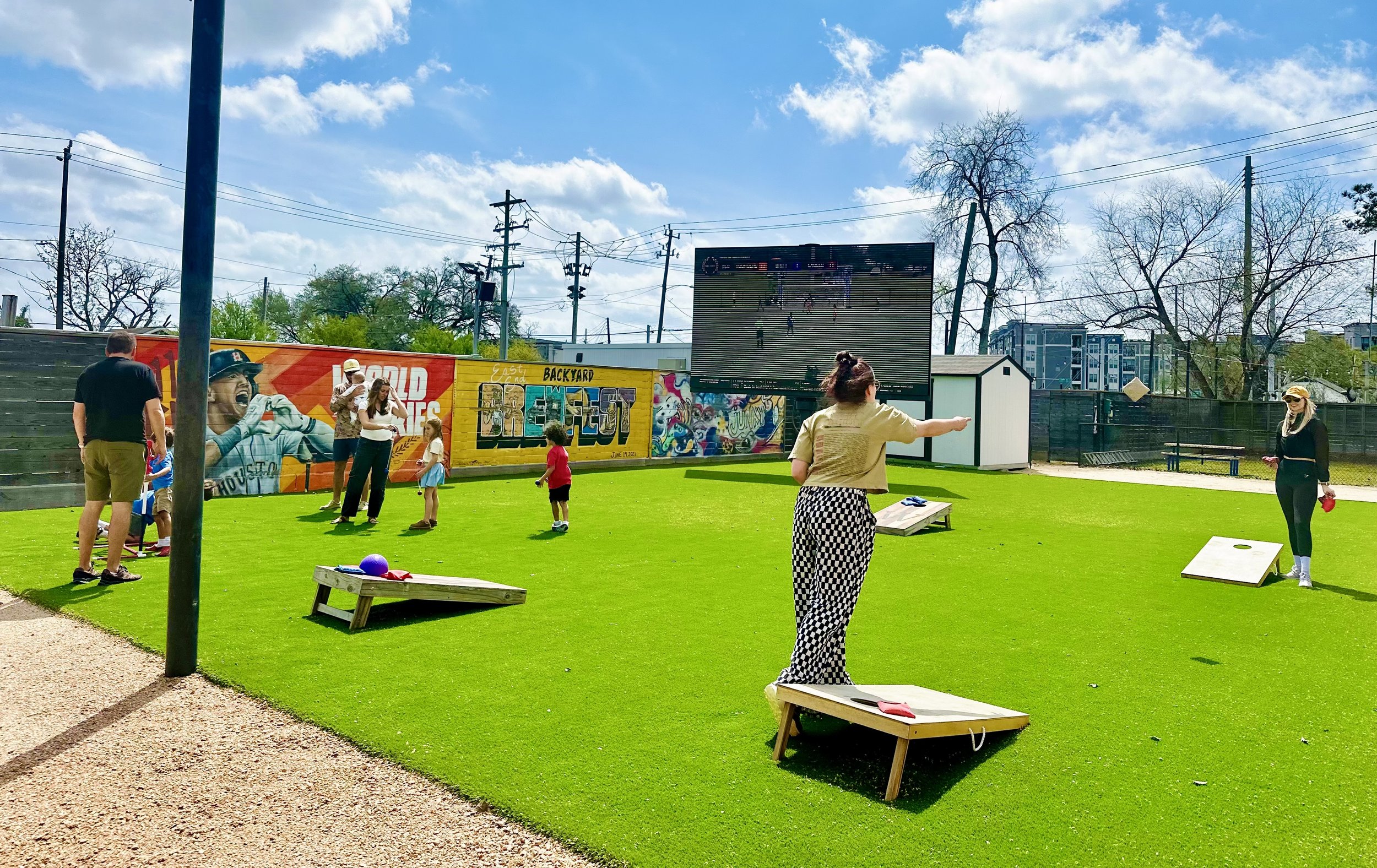 Group of people playing cornhole on a turf field at an outdoor event, with a mural and large screen in the background.