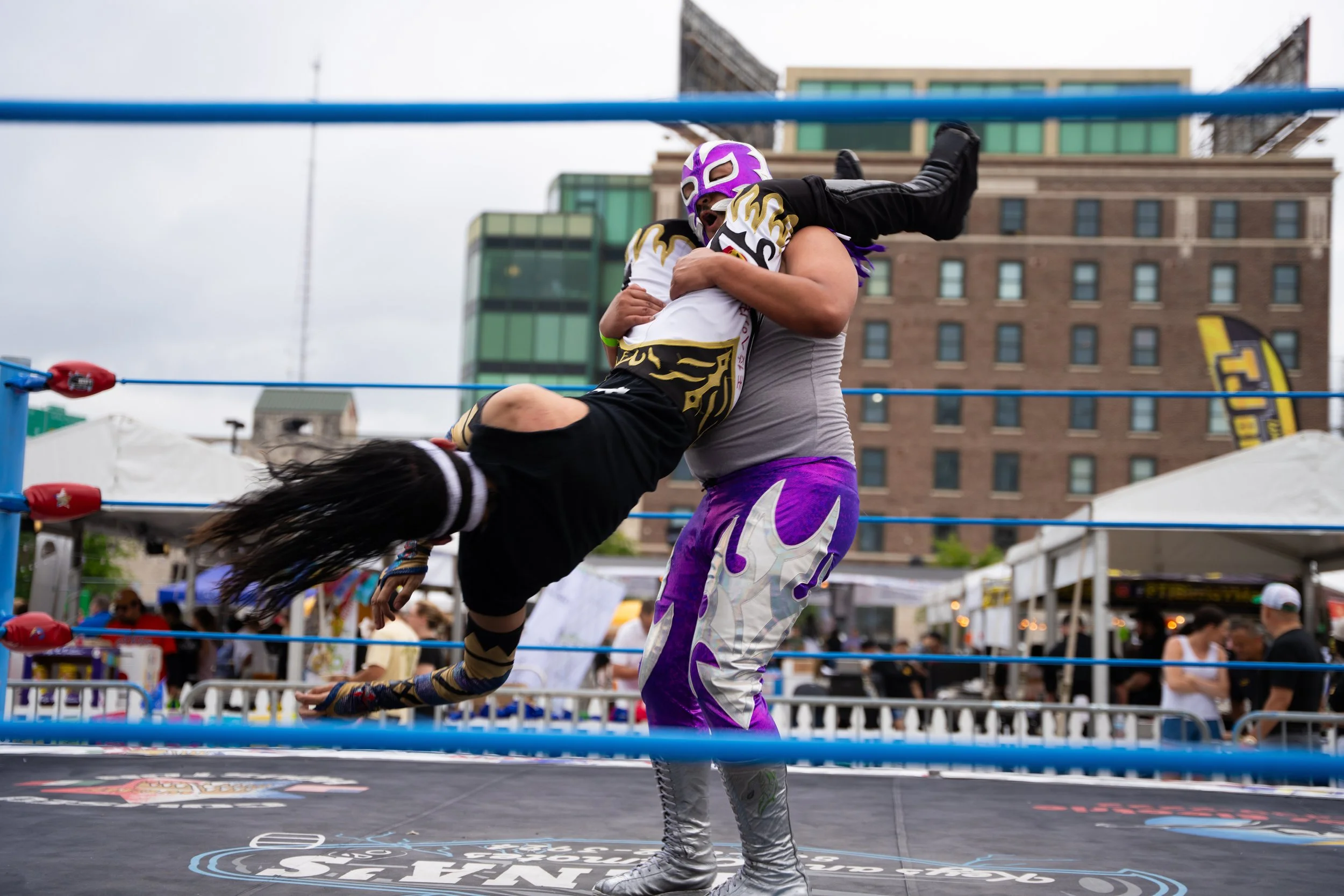 A professional wrestler in a purple mask and purple tights with silver designs lifts a young fan in a wrestling ring at an outdoor event.