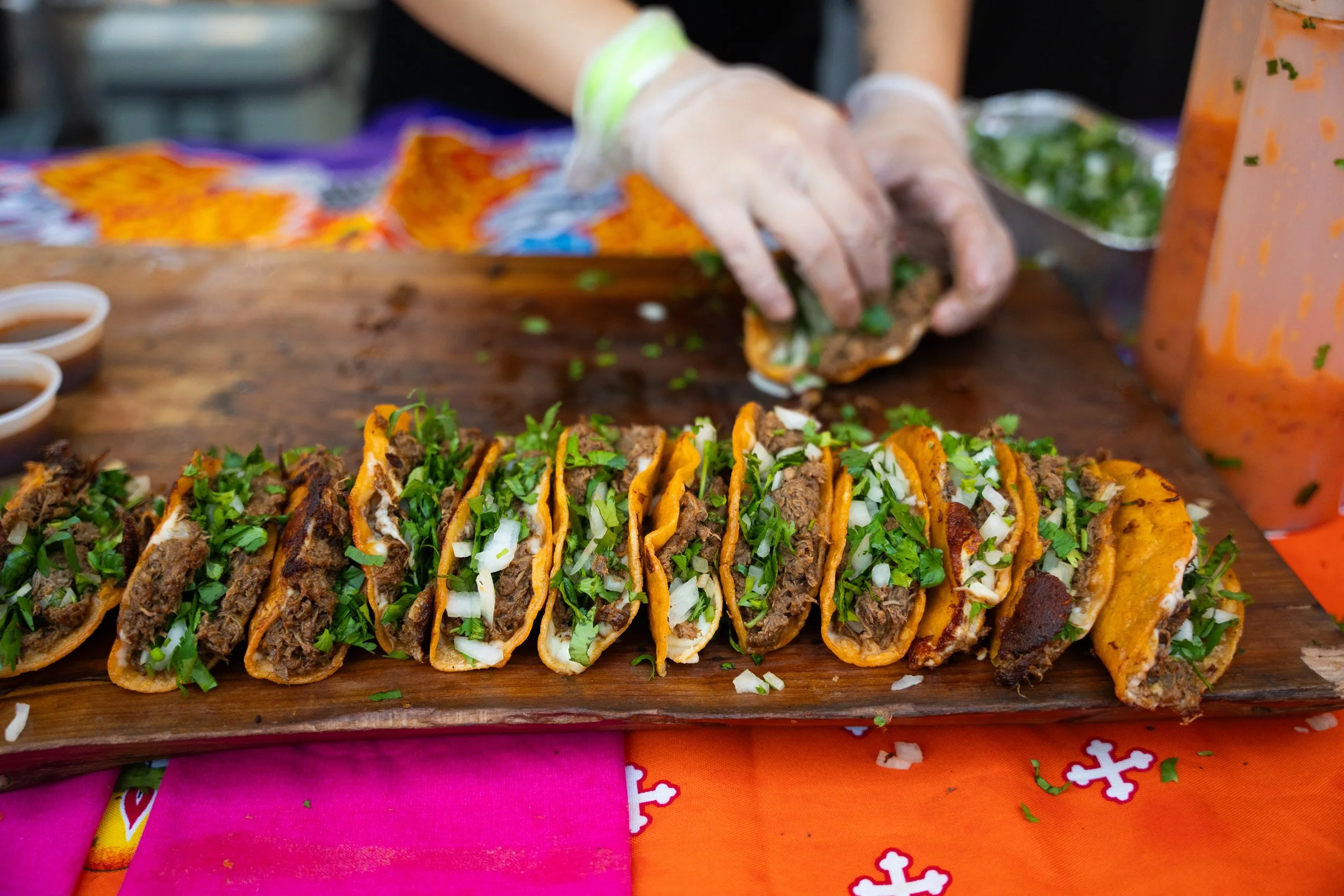 A row of Mexican tacos filled with beef, onions, and cilantro on a wooden serving board, with a person preparing another taco in the background.
