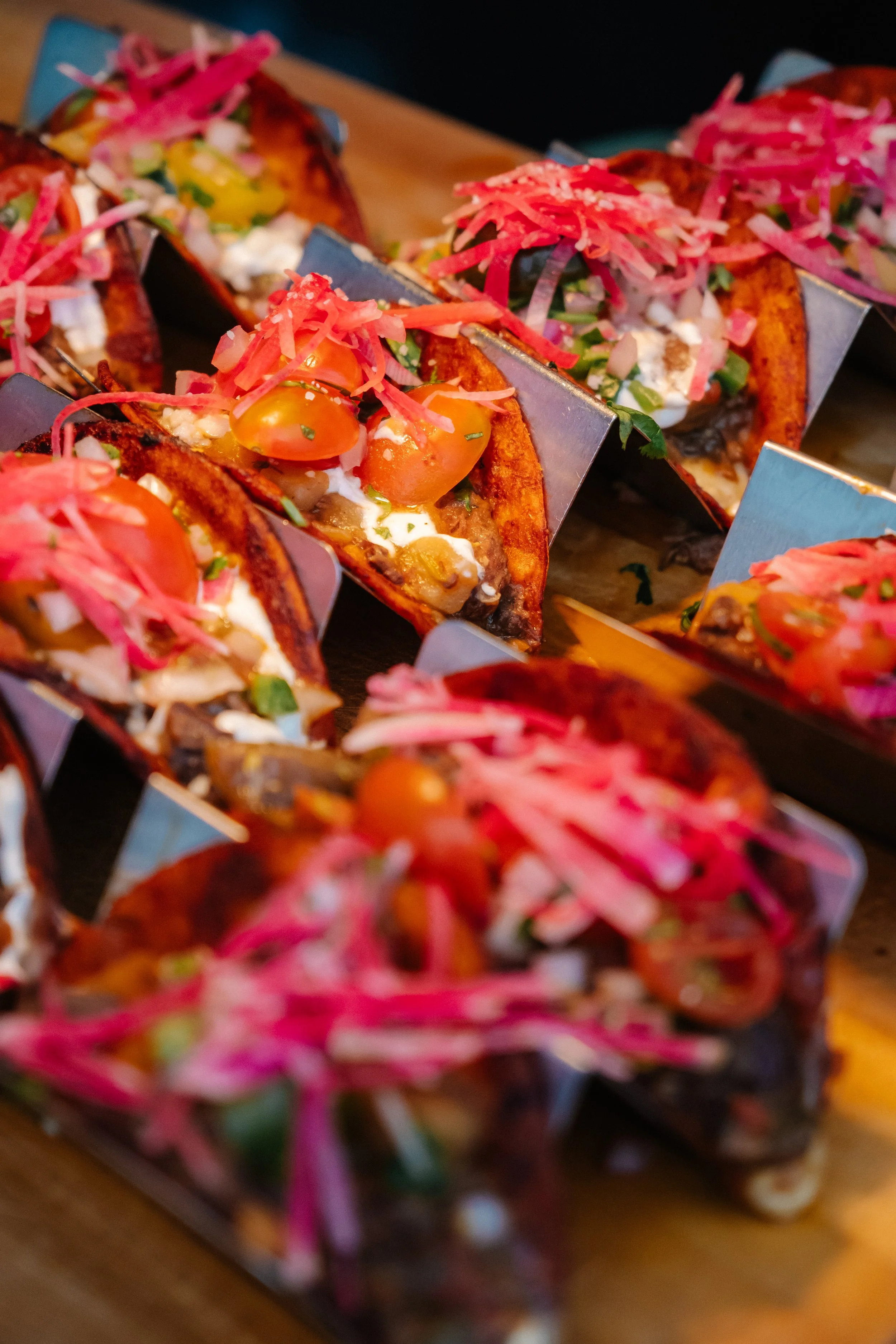 Close-up of several tacos topped with pink pickled onions, cherry tomatoes, sour cream, chopped green onions, and herbs.