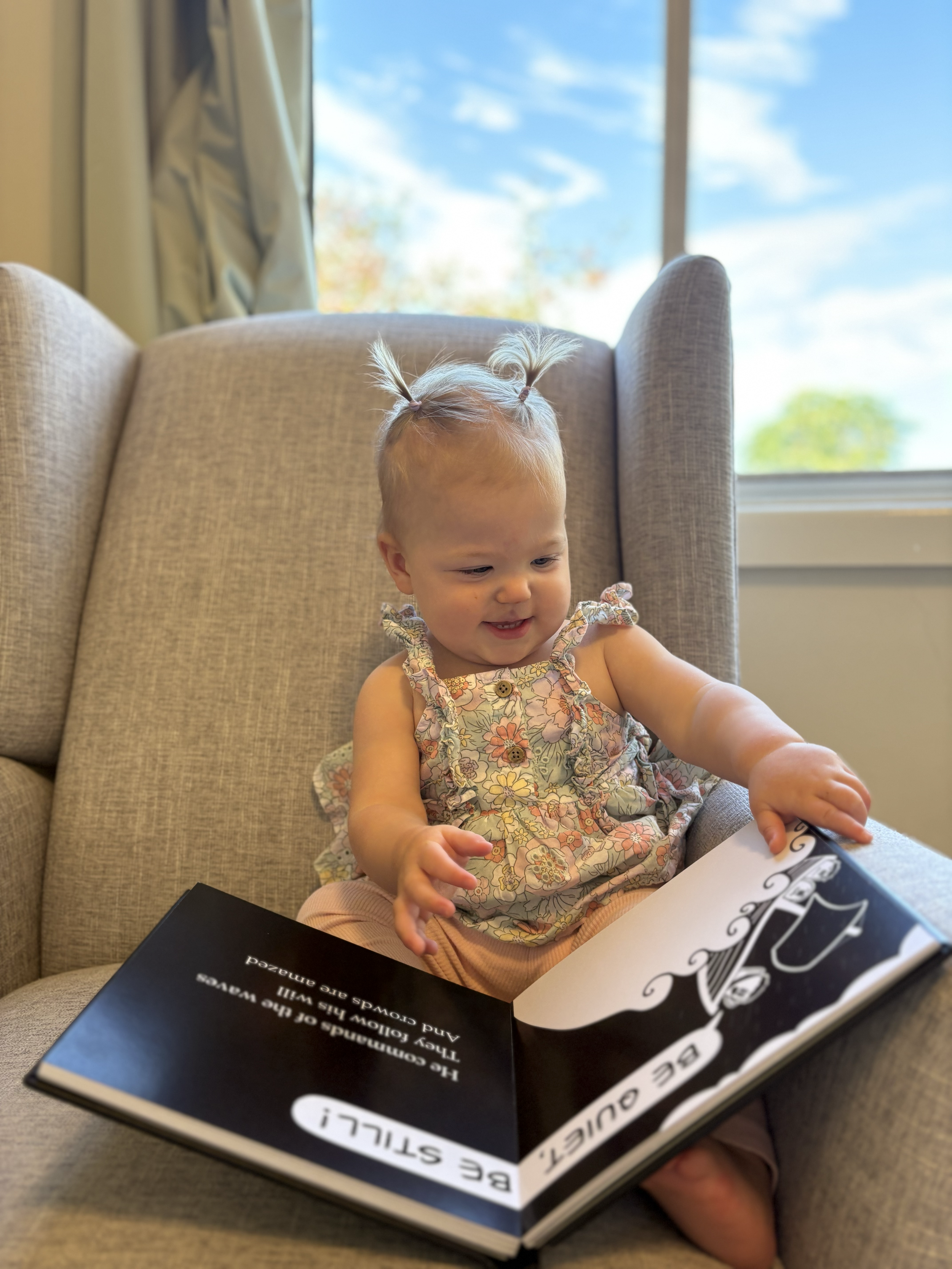 A young girl with blonde hair tied up in pigtails is sitting on a beige armchair, reading a black and white book. She is wearing a floral sleeveless top and light pink pants. There is a window with a view of blue sky and trees in the background.