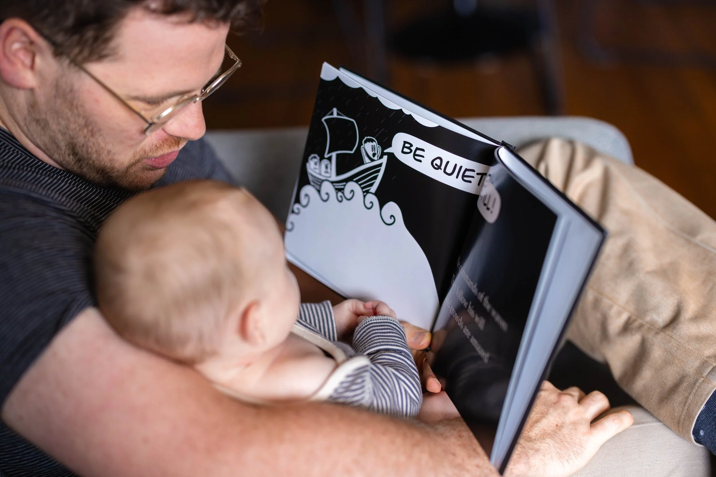 A man and a baby sitting on a bench, with the man holding a book or magazine that has a cartoon illustration of a boat and the words 'BE QUIET' on the cover.