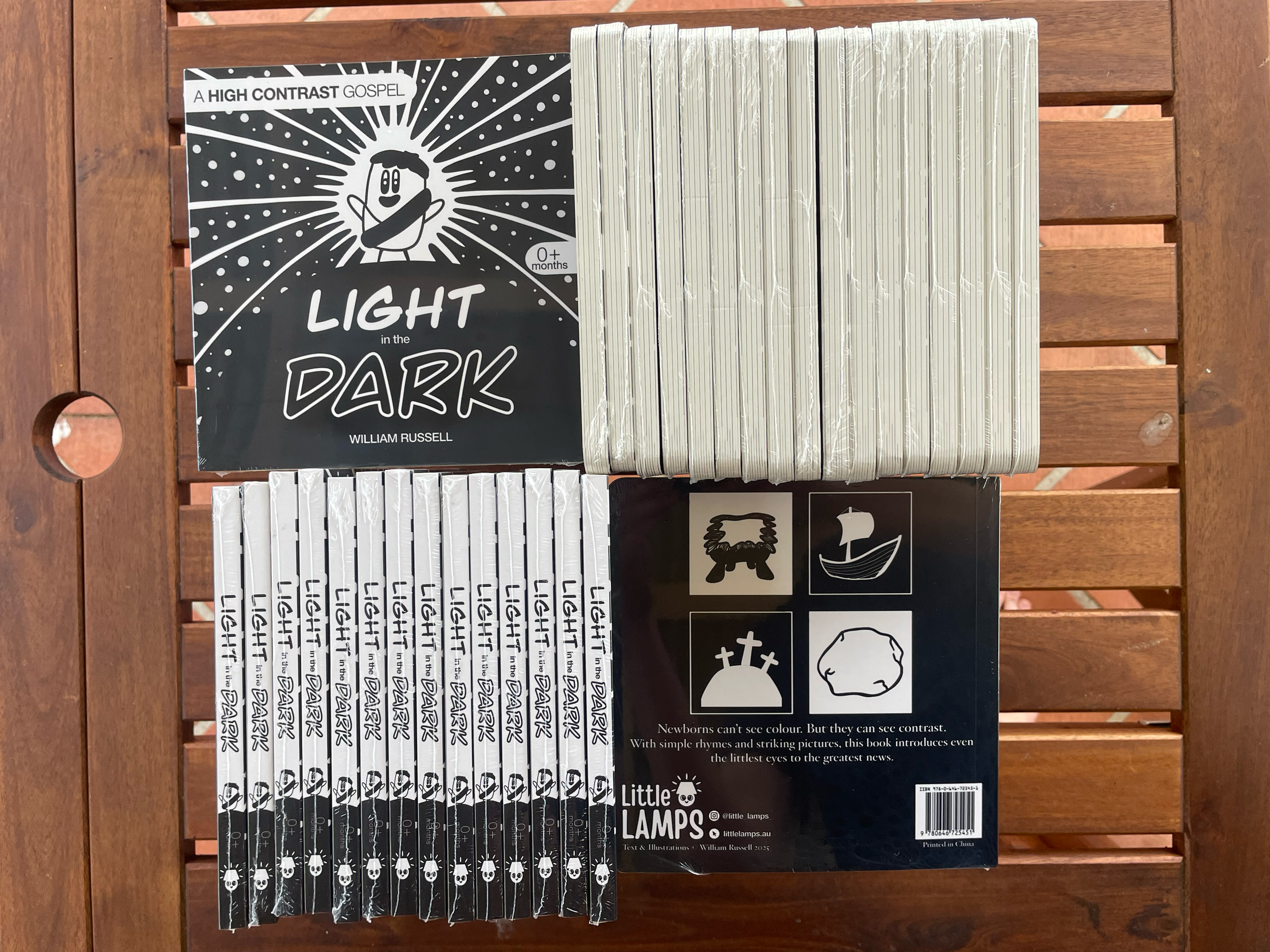 Books titled 'Light in the Dark' by William Russell, with black and white covers featuring a cartoon character, and a black and white box with symbols of a lamb, a sailboat, a grave, and a rock, on a wooden surface.