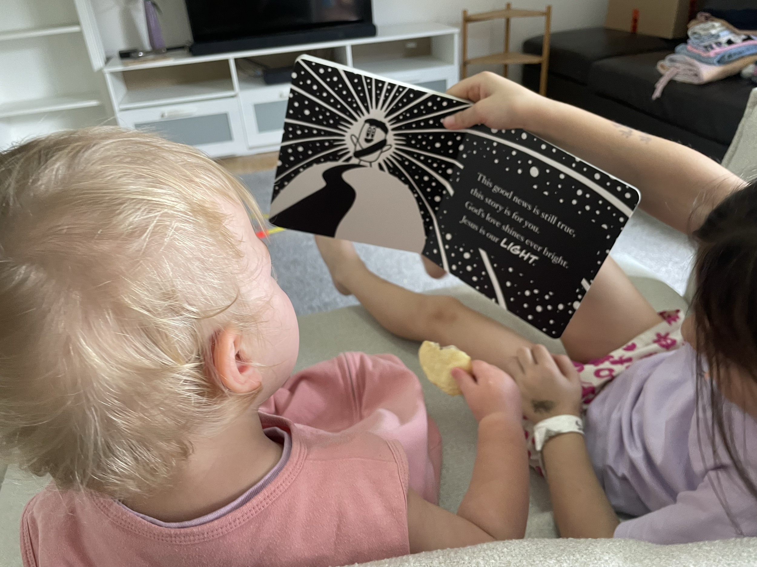 Two children sitting on a couch, one with blonde hair and the other with darker hair, looking at a black and white illustrated card with a quote. The card features a stylized image of a smiling face with rays, a winding path, and text that reads, 'Th