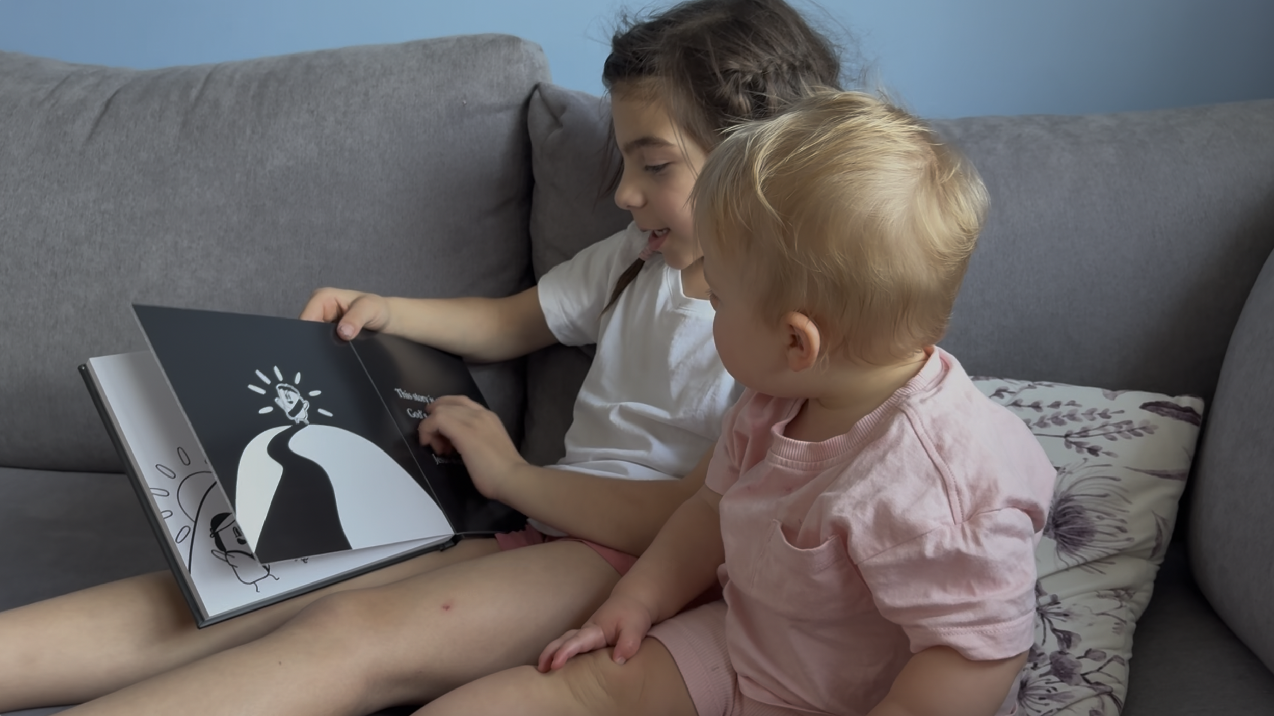 Two young girls, one older with dark hair and one younger with blonde hair, sitting on a grey couch, looking at a children's picture book called 'Light in the Dark' together.