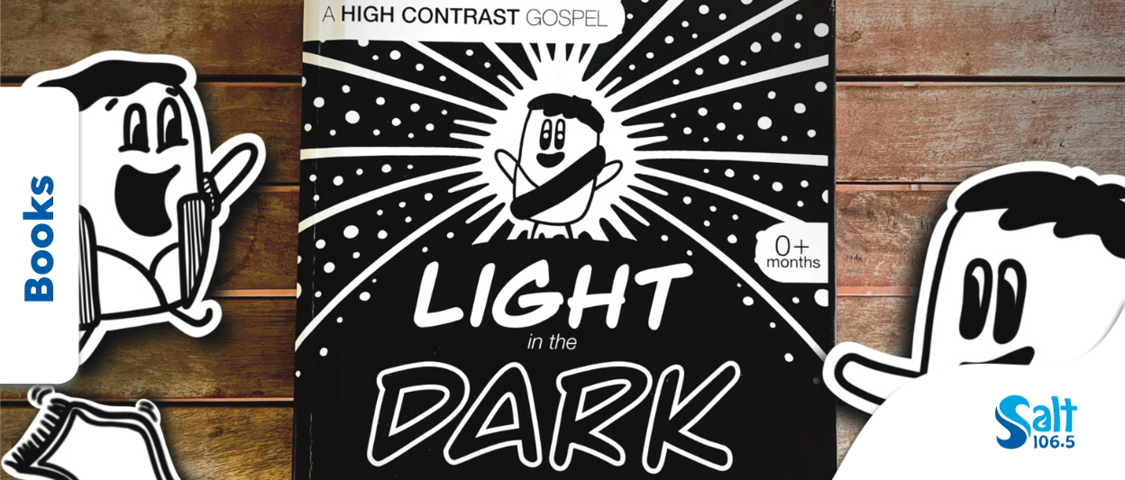 Cover of a children's book titled 'Light in the Dark' with a cartoon character surrounded by radiating lines and stars, indicating it is suitable for ages 0 months and up, displayed on a wooden surface.