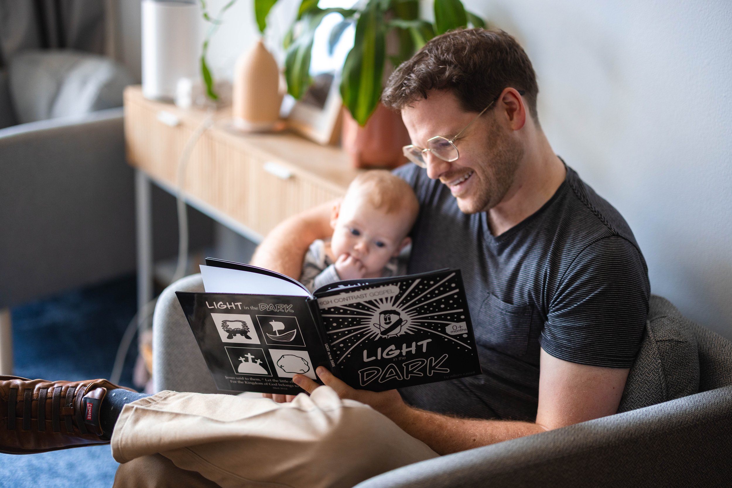 A man with glasses and a beard reading a children's book titled "Light in the Dark" to a baby sitting on his lap in a cozy living room with plants and furniture.