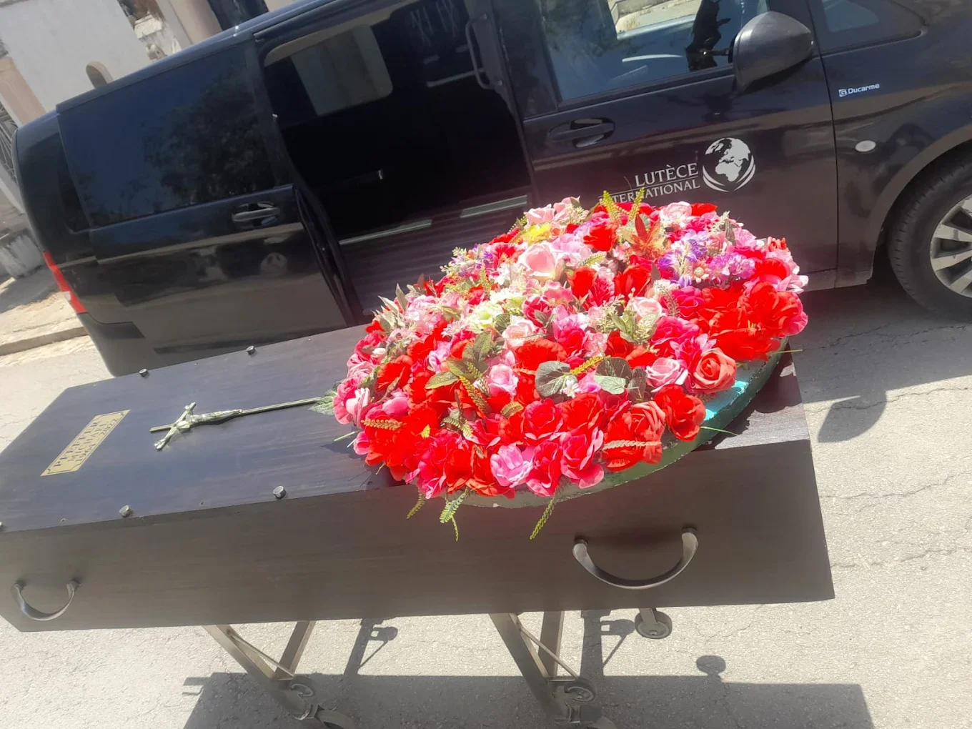 Une bière en bois avec un crucifix et un bouquet de fleurs colorées, situé à côté d'une voiture noire portant un logo de lutèce international.