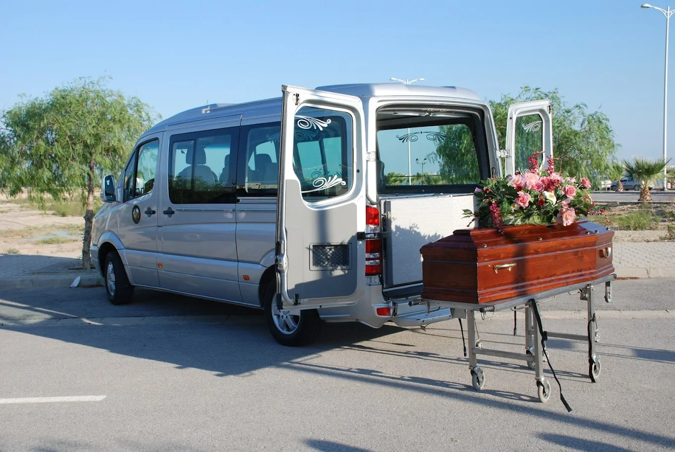 Une voiture funéraire blanche avec une caisse en bois et des fleurs sur un chariot à roulettes dans un parking en plein air, avec des arbres en arrière-plan.