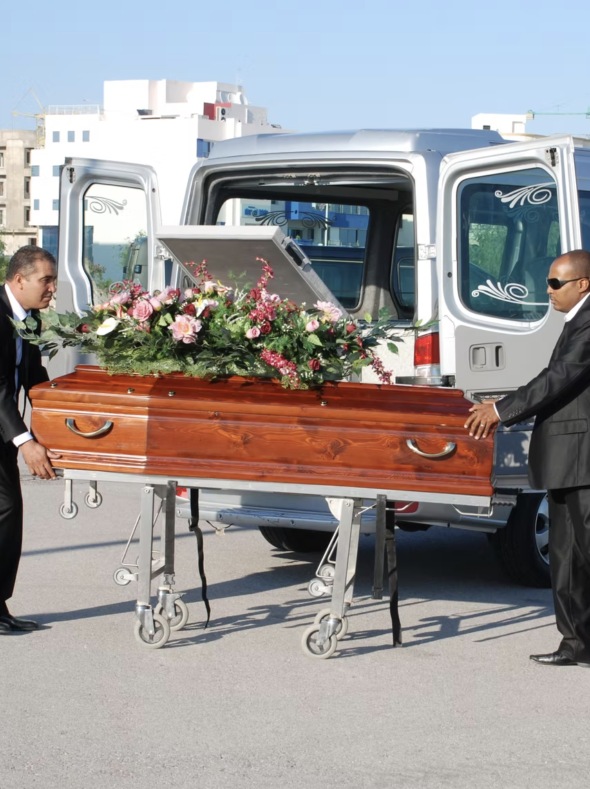Deux hommes en costumes noirs transportent un cercueil en bois décoré de fleurs dans une camionnette ouverte, apparemment lors d'une cérémonie funéraire en plein air.
