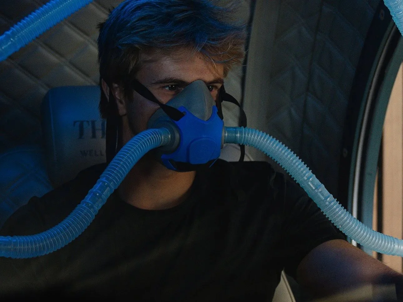 A person wearing a protective oxygen mask connected to a blue and gray breathing apparatus, sitting in a confined space.