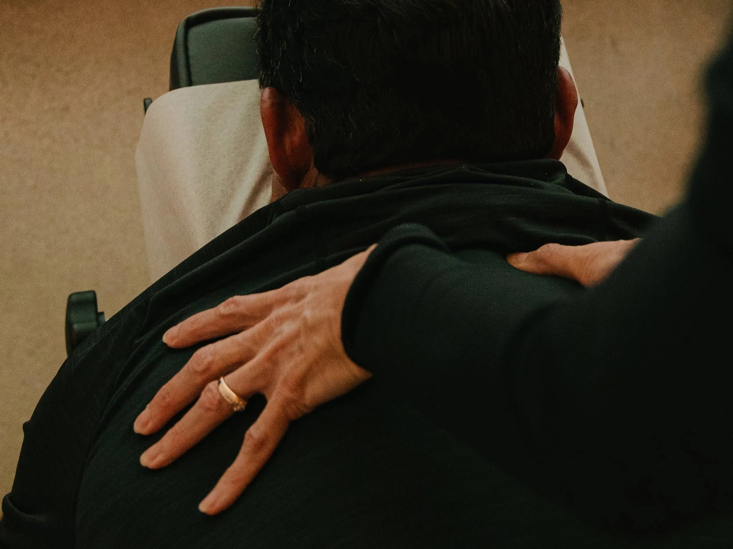 A person's back as they sit in a chair, with a hand resting on their shoulder, wearing a dark top.