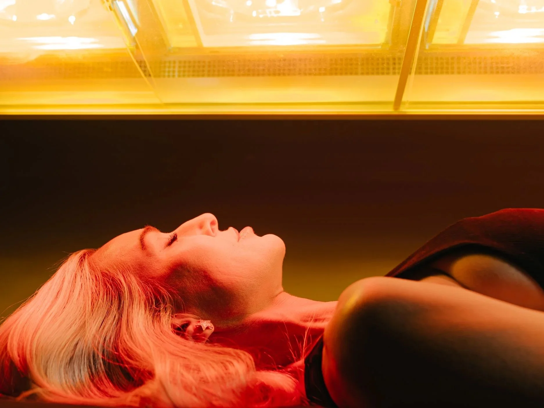 Close-up of a woman with long, wavy blonde hair lying down, eyes closed, under warm yellow lighting.