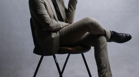 Executive men dressed in a beige suit sitting on a chair with crossed legs and one hand resting on their face. confident men, how to be a great lover?, how to be a better lover?