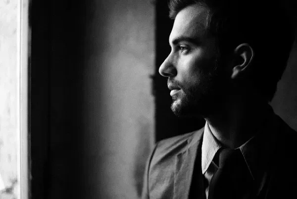 A black and white profile of a man looking out a window, wearing a suit and tie.