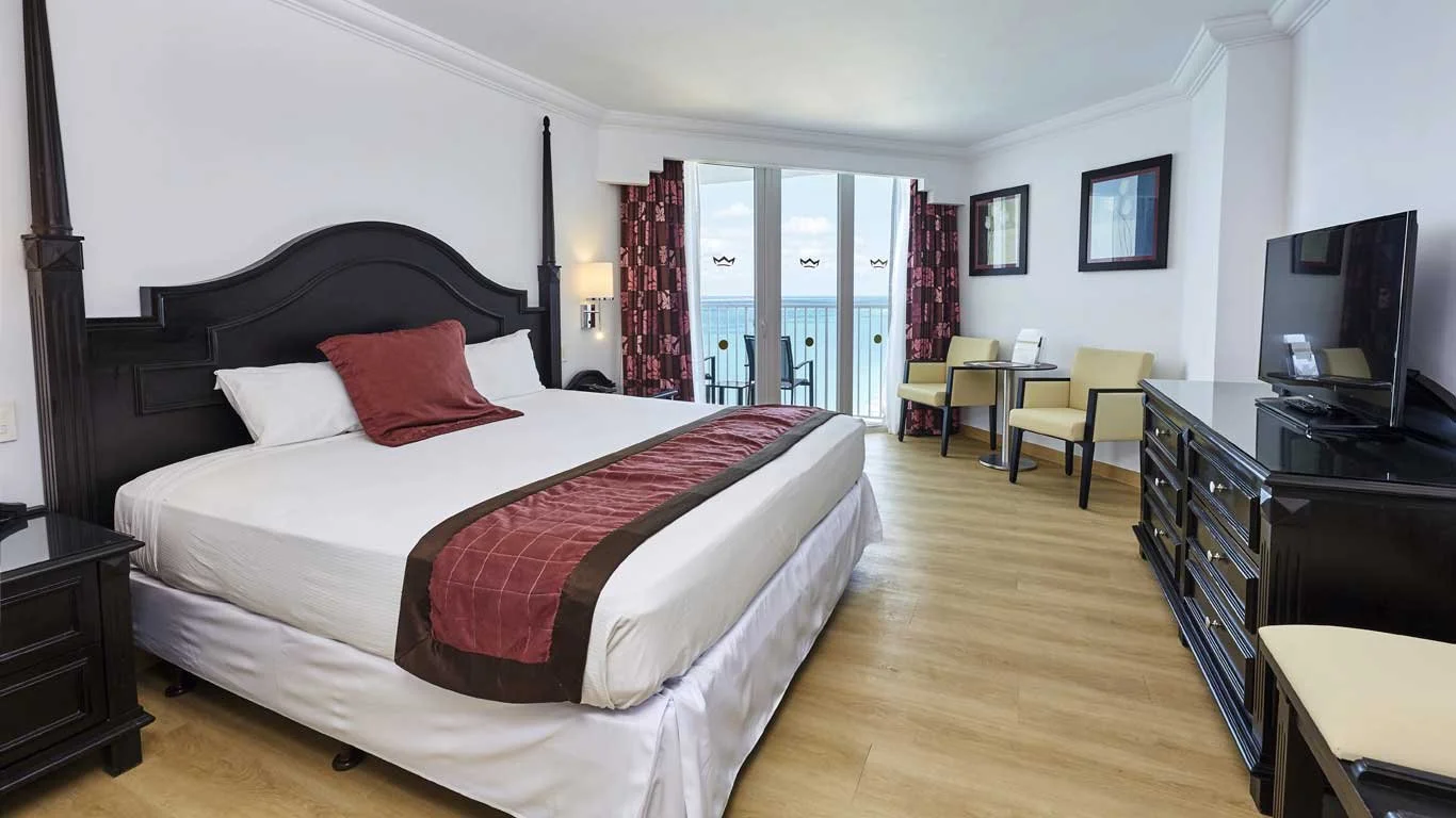 A hotel room with a large bed, black headboard, white bedding, a red and brown bed runner, and a single red pillow. There are two chairs and a small table near a sliding glass door with a balcony overlooking the ocean. The room has a black dresser with a TV, framed artwork on the walls, and light wooden flooring.