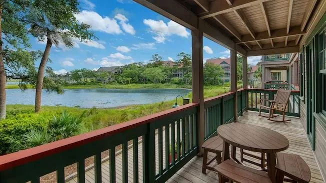 Balcony overlooking a pond with trees and houses in the background, wooden railing and furniture.