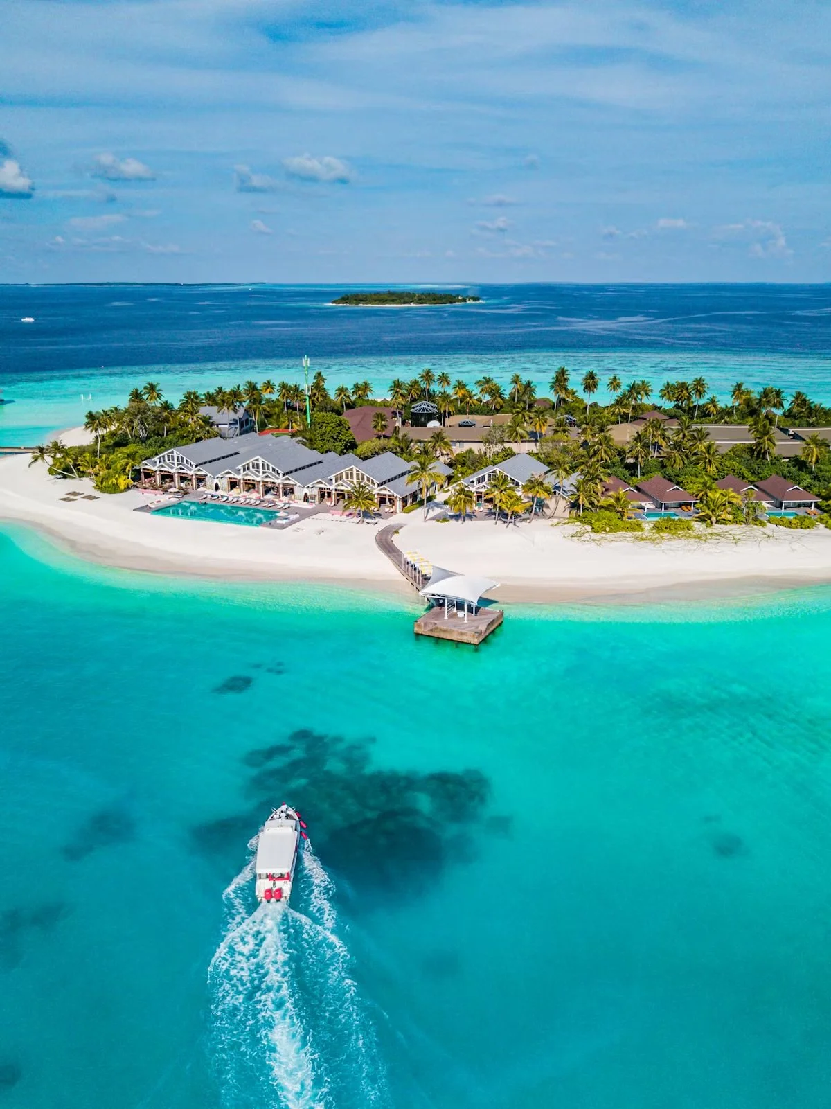 Aerial view of a tropical island with white sandy beaches, turquoise waters, palm trees, a resort with buildings and a swimming pool, and a boat heading towards the island.