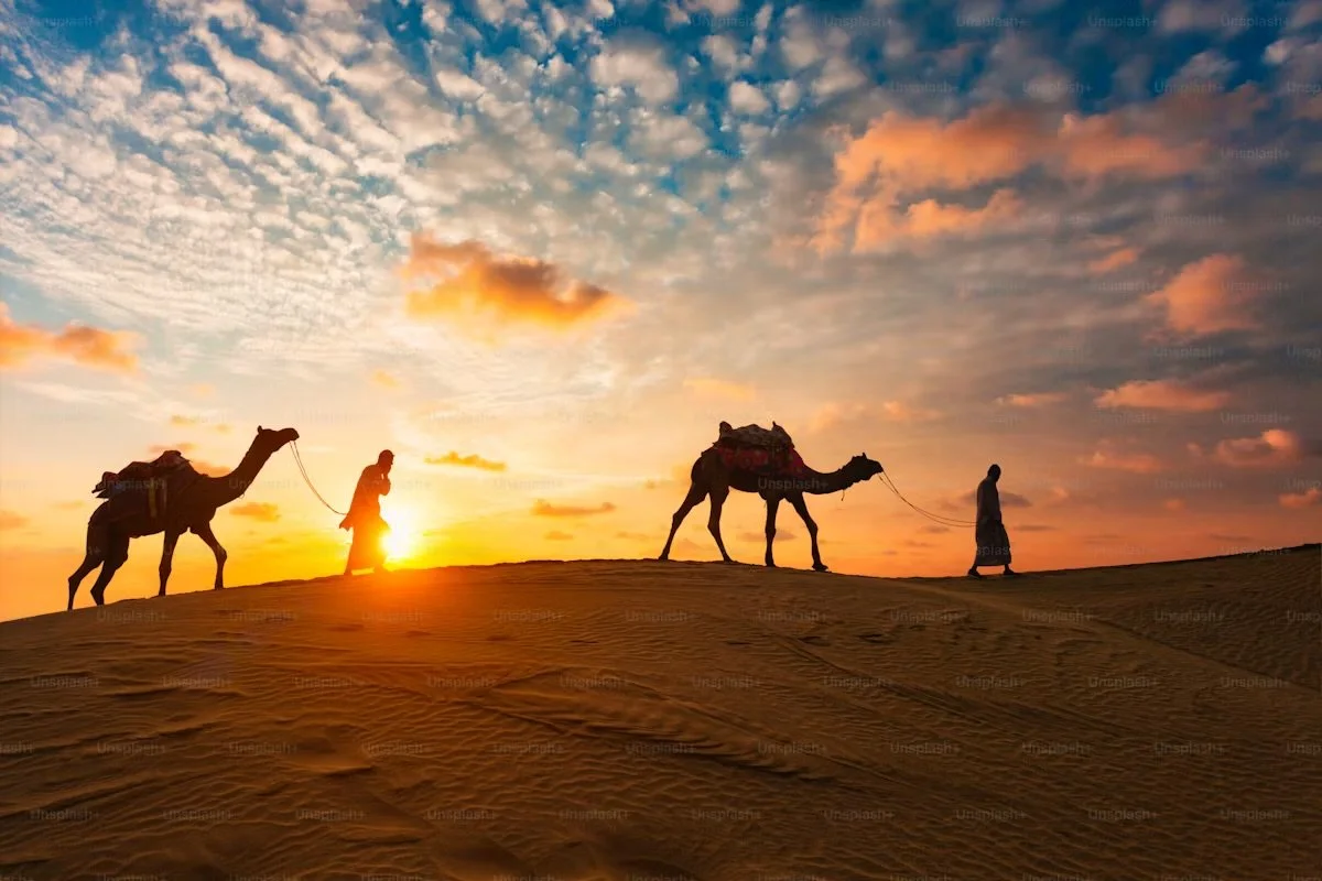 Silhouettes of two men walking with camels in a desert at sunset, with orange and blue sky and scattered clouds.