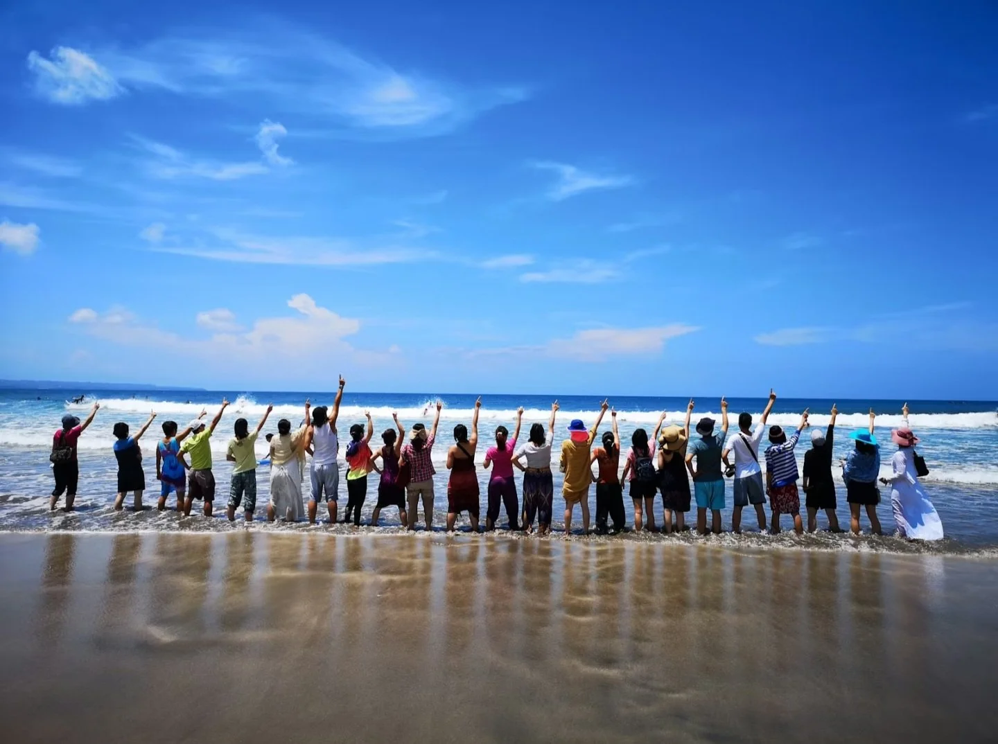 Group of people standing on the beach, facing the ocean, with their hands raised, under a partly cloudy blue sky.