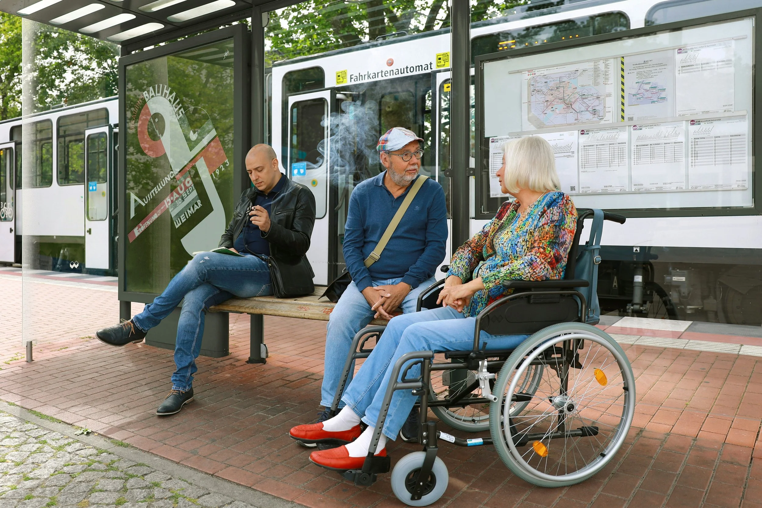 Three people sitting at a bus stop with a bus in the background. One man is looking at his phone, a woman in a wheelchair is talking to another man.