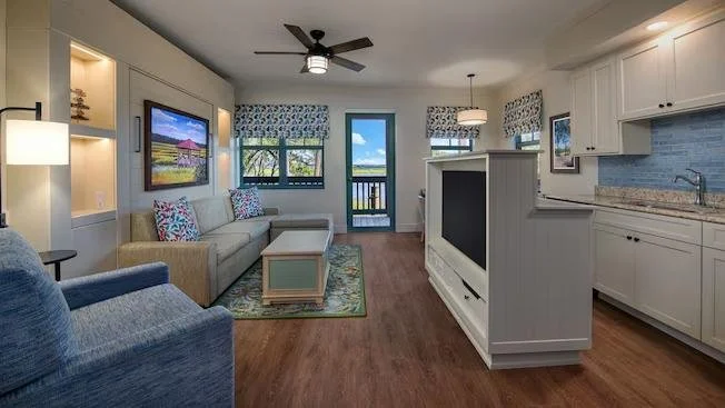 Living room and kitchen area with modern decor, a sofa, armchair, flat-screen TV, windows with valances, and a kitchen with white cabinets and granite countertops.