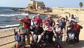 A group of elderly people in wheelchairs and walkers on a seaside walkway with a historic stone fortress and ocean in the background.