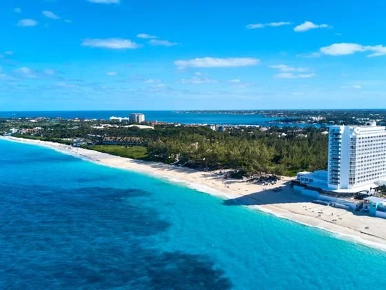 Aerial view of a coastal city with a beach, ocean, and high-rise buildings under a blue sky with scattered clouds.