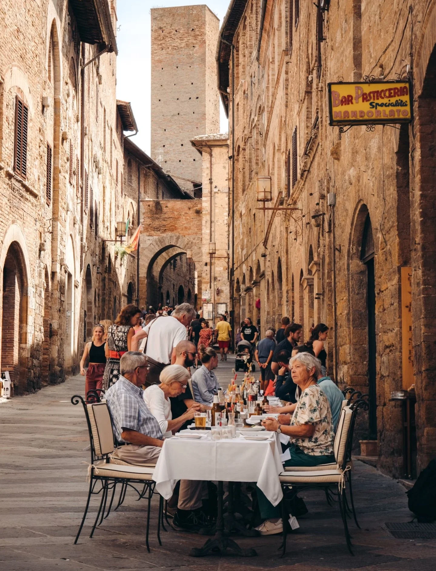 People dining at an outdoor restaurant on a narrow cobblestone street in an old European city with tall brick buildings and a tower in the background.