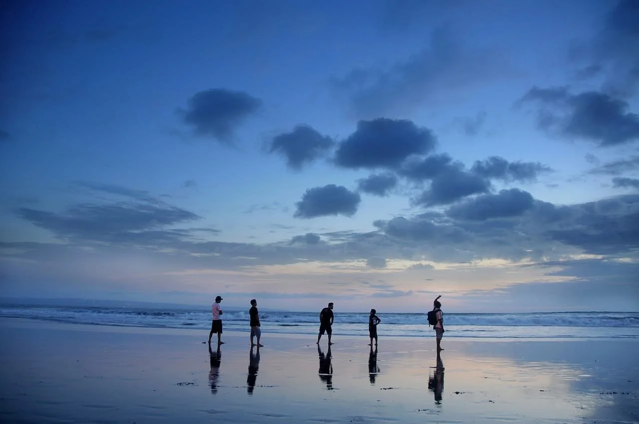 Five people walking along the beach with ocean waves and cloudy sky during sunset or sunrise.