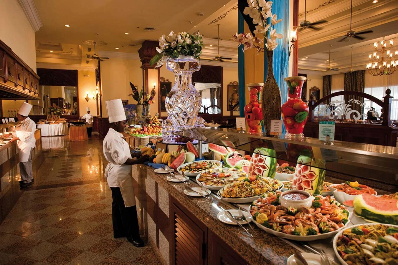 A buffet restaurant with a variety of colorful foods and desserts, including fruits, salads, and seafood, in a warmly lit elegant dining space with chefs in white uniforms and tall chef hats