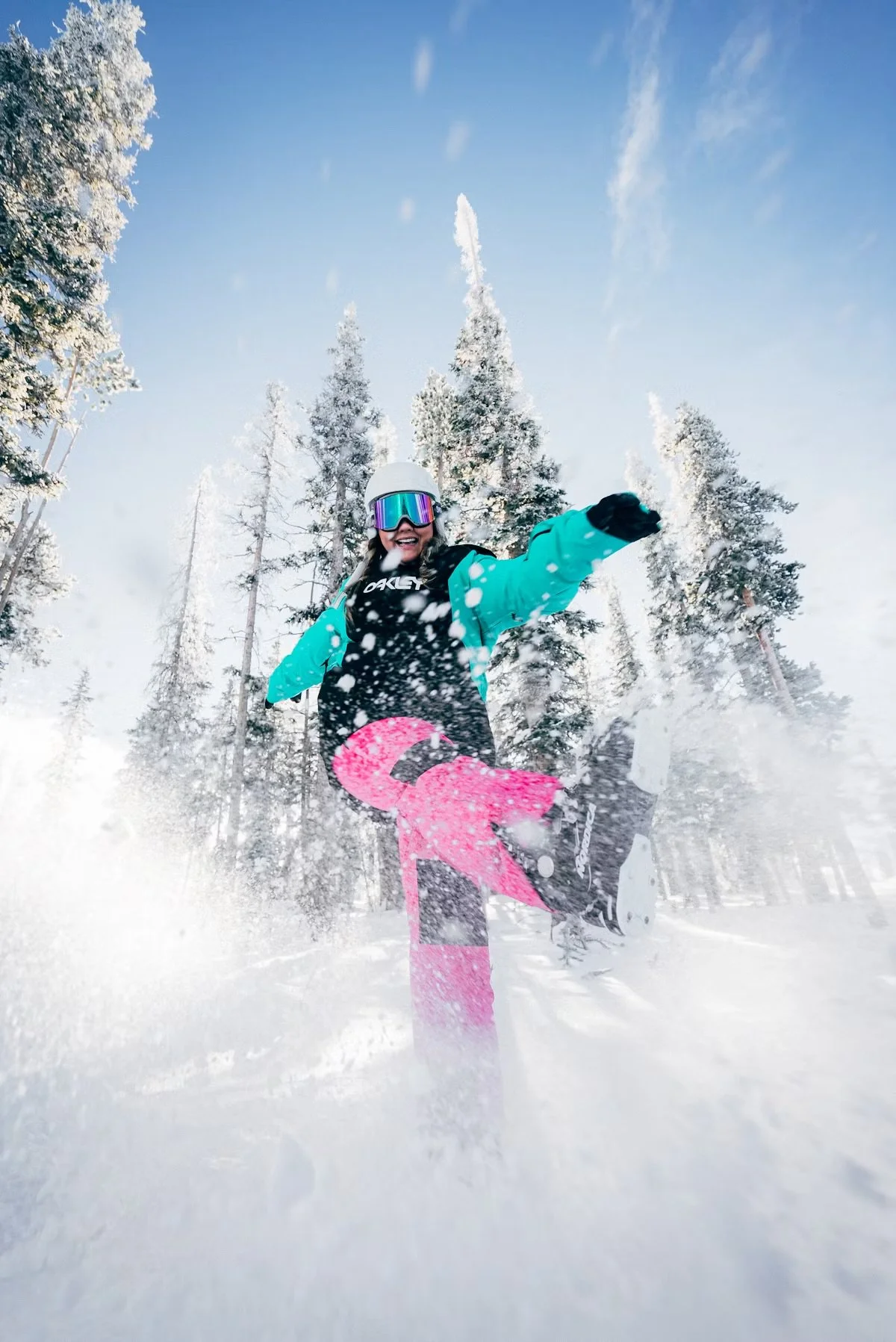Child snowboarding in a snowy forest, wearing a blue jacket, pink snow pants, white helmet, and ski goggles, kicking up snow under a clear blue sky.