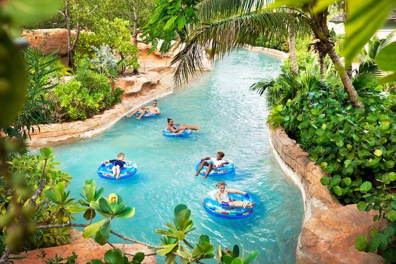 People relaxing on inner tubes in a lazy river at a water park surrounded by lush green tropical plants and trees.