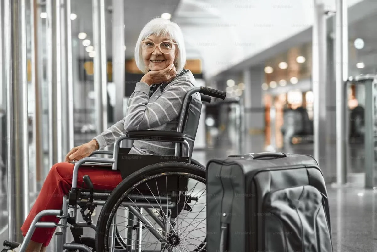 An elderly woman with white hair and glasses sitting in a wheelchair at an airport, smiling, with a black suitcase nearby.