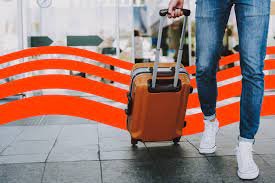Person walking through airport with brown rolling suitcase, wearing jeans and white sneakers.