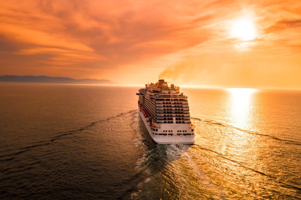 A large cruise ship sailing on the ocean at sunset with a vibrant, orange sky and the sun reflecting on the water.