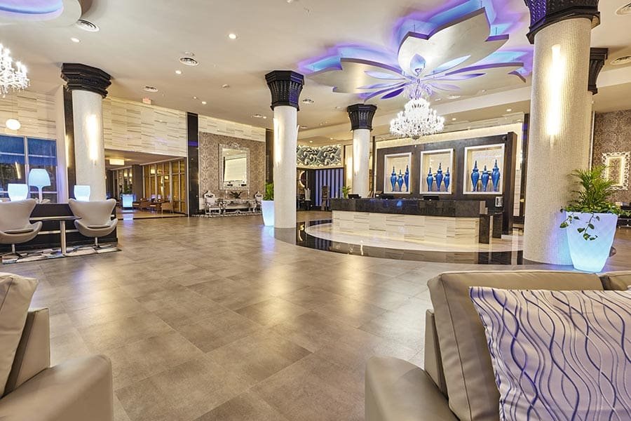 A luxurious hotel lobby with a spacious open area, elegant chandeliers, decorative columns, modern furniture, a curved reception desk with blue vases artwork, and ambient lighting.