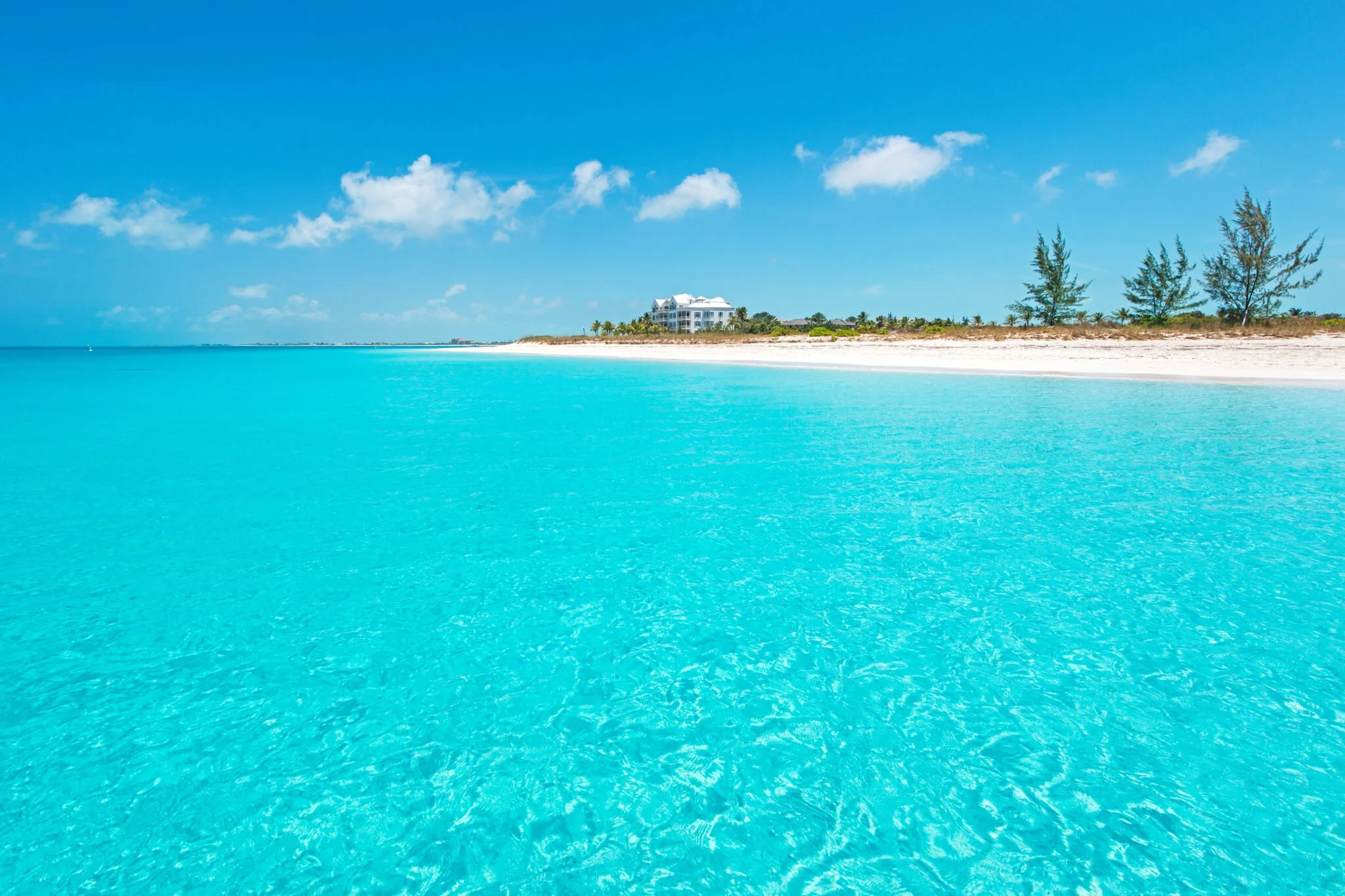 Clear turquoise ocean water along white sandy beach with a few trees and a large building in the distance.