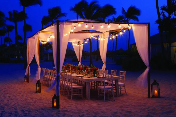 Outdoor beach dinner setup under a canopy with string lights, a long table, chairs, and lanterns, with palm trees in the background at dusk.