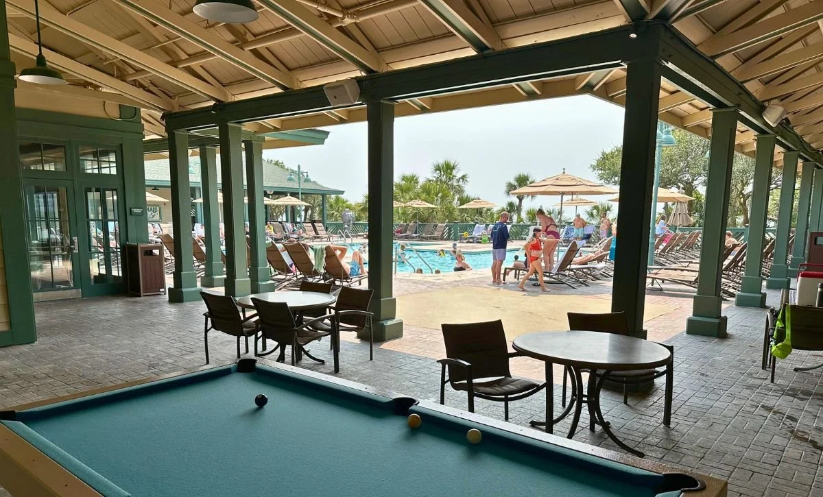 View from inside a poolside cabana looking out onto a swimming pool area with lounge chairs, umbrellas, and people enjoying the pool.