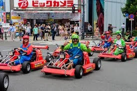 People riding go-karts on a city street with a shopping mall in the background.