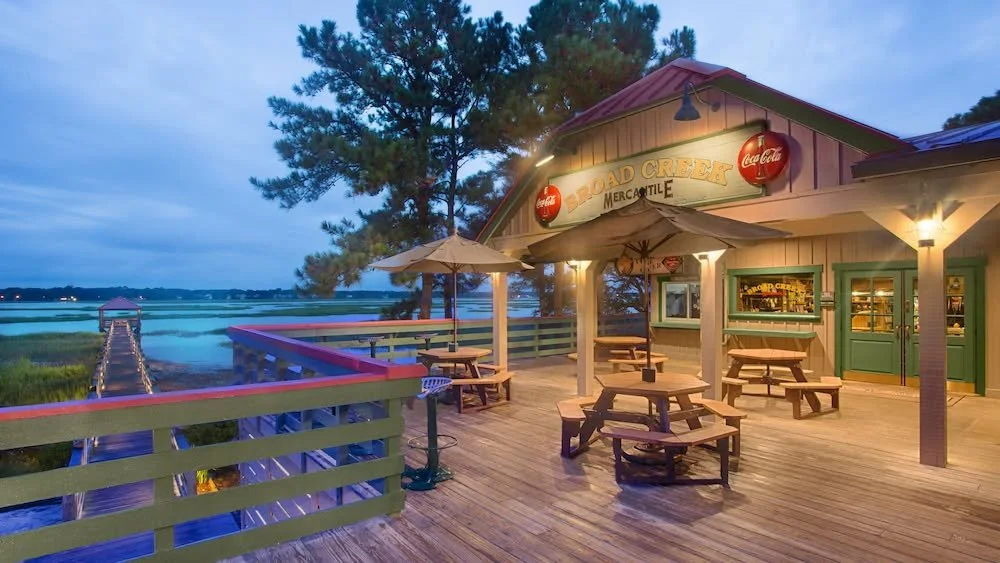 Outdoor dining area at Road Creek Mercantile with picnic tables, umbrellas, and a view of a salt marsh and boardwalk during dusk.