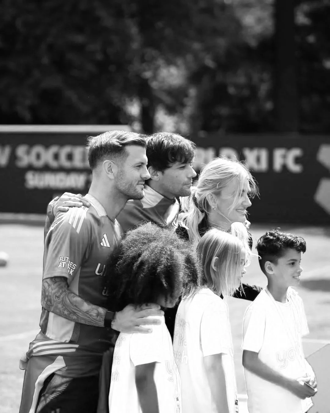 A black and white photo of two adult males, a woman, and three children standing outdoors on a sports field, with a blurred sign behind them. The group is smiling and looking towards the right.